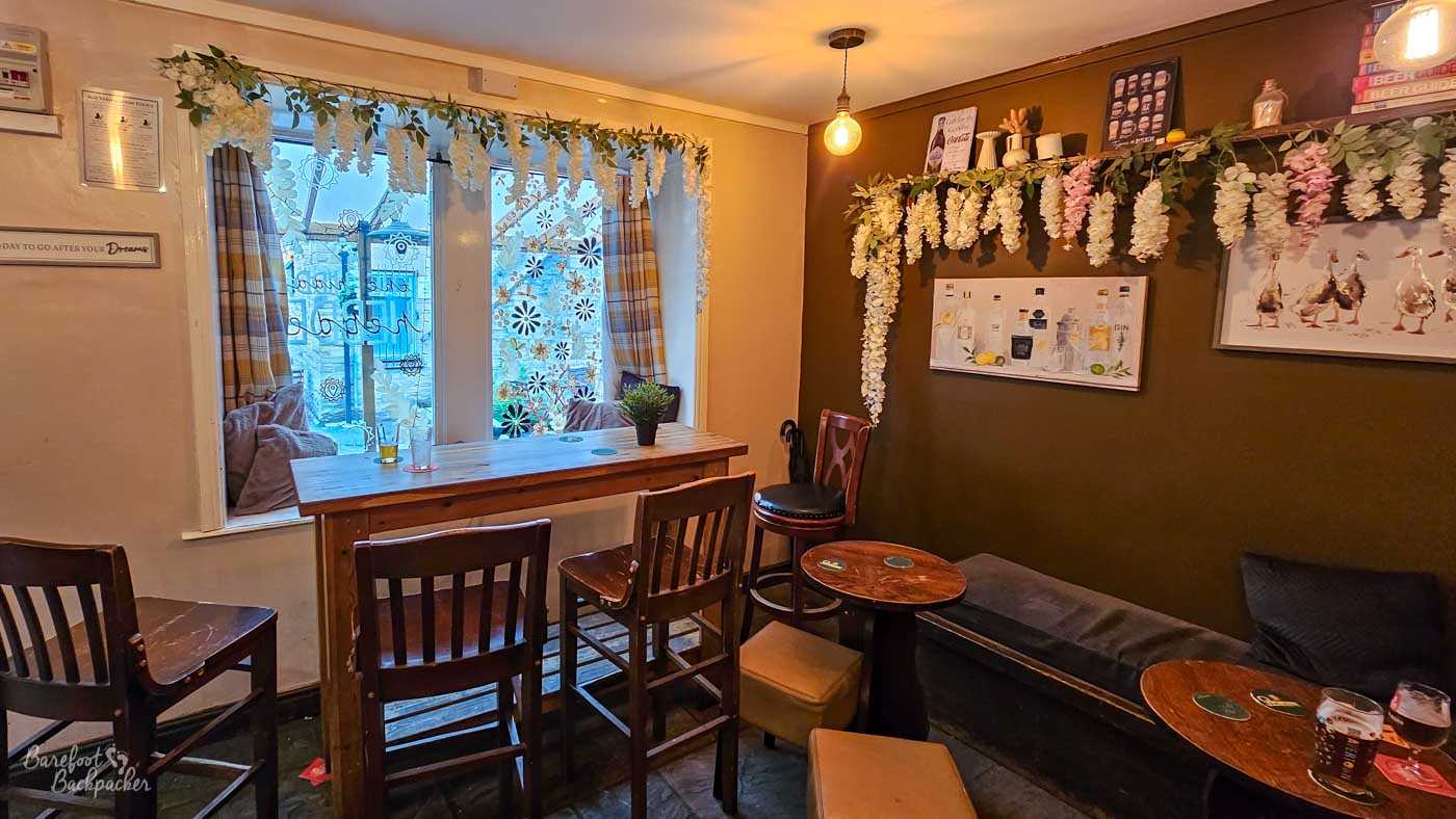 Cosy seating area inside The Hidden Hebden in Hebden Bridge, with dark brown walls, wooden furniture and a long cushioned bench along one side. A high table sits across a window decorated with floral patterns, while artificial white flowers and trailing greenery frame both the window and walls. Gin-themed artwork and a picture of geese hang among the decorations, and warm bare-bulb lighting adds to the intimate atmosphere.