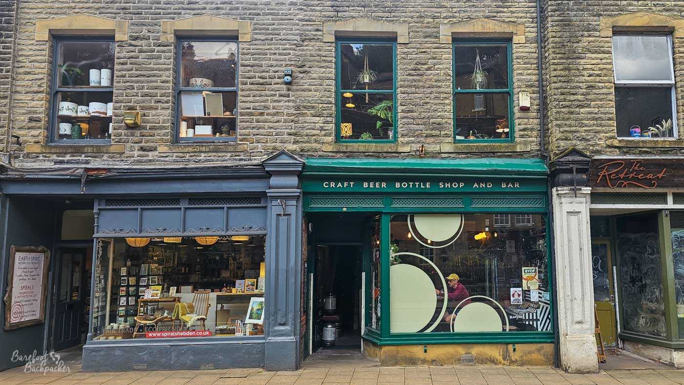 Street frontage of Drink?, a craft beer bottle shop and bar in central Hebden Bridge, although the bar’s name itself is not displayed outside. Its dark green shopfront carries gold lettering reading CRAFT BEER BOTTLE SHOP AND BAR, with large cream circles decorating the window and shelves of bottles visible inside. It occupies the ground floor of a traditional stone building between an art and eco shop on the left and The Retreat on the right, with upper-storey sash windows above.