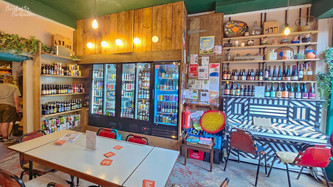 Bright, eclectic interior of Drink? in Hebden Bridge, with a large communal table and mismatched chairs in the foreground. Wooden shelves and three tall illuminated fridges are packed with colourful cans and bottles of craft beer, while more bottles, including cider, line shelves on the right. A black-and-white geometric upright piano stands against the wall alongside plants, pottery, notices and a circular POPUP TODAY sign. Bare bulbs and timber panelling add to the informal, quirky bottle-shop atmosphere.