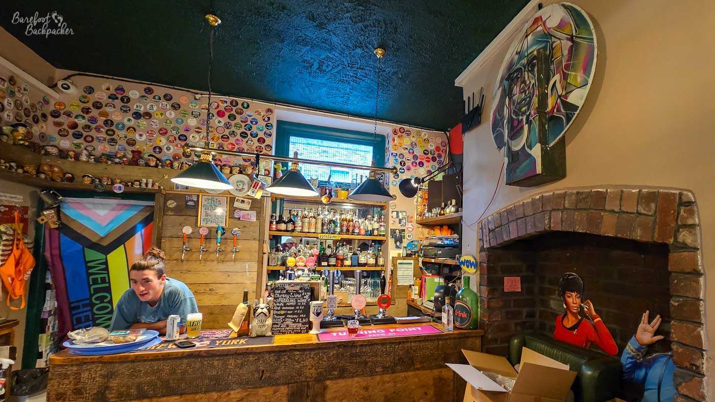 The small, colourful bar at Drink? in Hebden Bridge, packed with quirky decoration. Beer taps and bottles crowd the wooden bar beneath three hanging lamps, while dozens of colourful beer badges cover the upper walls and a large Progress Pride flag hangs to the left. On the right, a brick fireplace contains a life-size image of Star Trek character Uhura sitting in an armchair, beneath a brightly coloured portrait. The dark green ceiling and eclectic collection of objects give the room an intimate, playful feel.