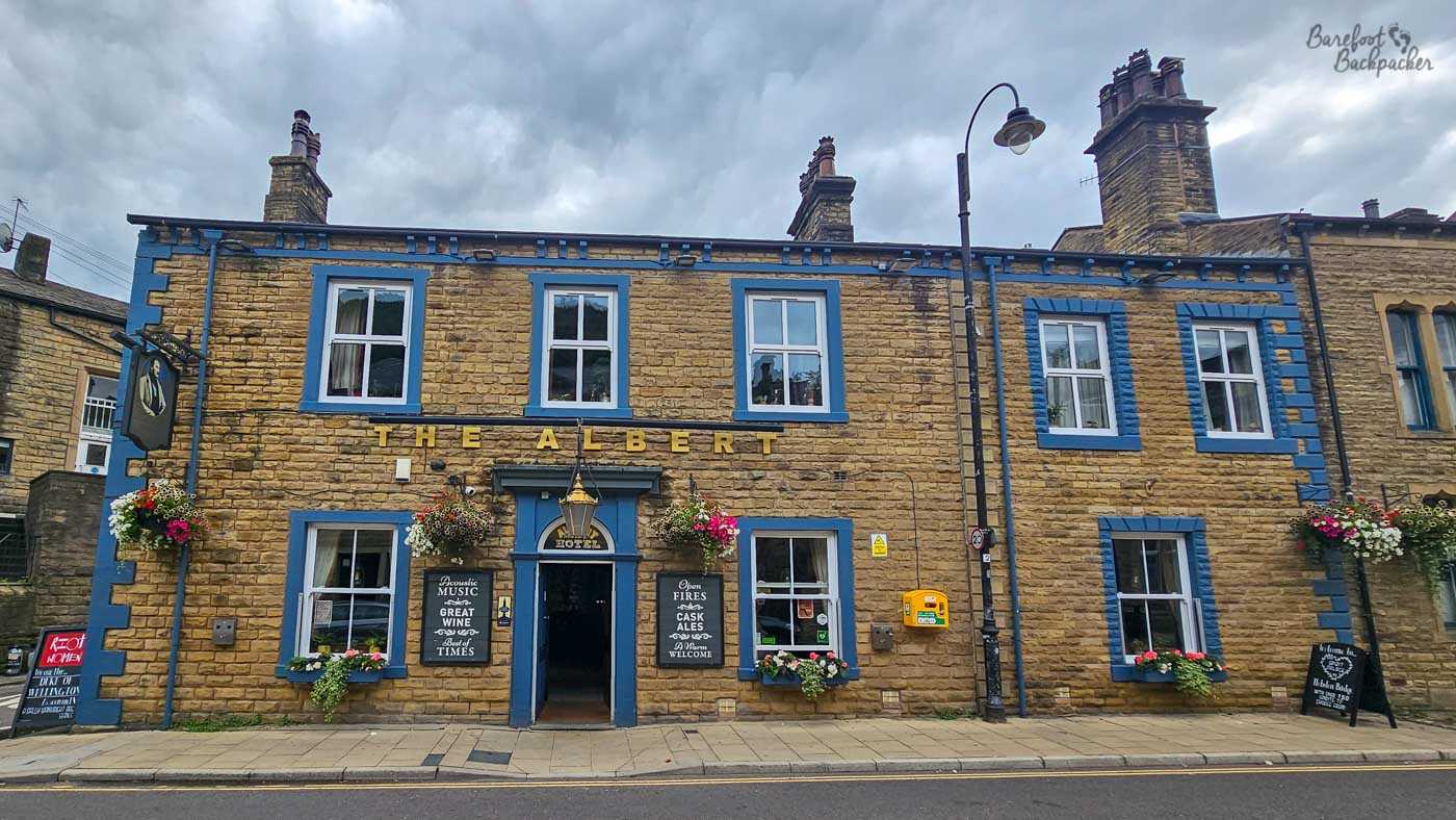 Exterior of The Albert in Hebden Bridge, a substantial two-storey traditional stone pub with distinctive blue-painted trim around its white-framed windows, corners and roofline. Gold letters spelling THE ALBERT stretch across the frontage above the central entrance, which is flanked by blackboards advertising music, wine, fires and cask ales. Colourful hanging baskets and window boxes brighten the honey-coloured stonework beneath a heavily overcast sky.