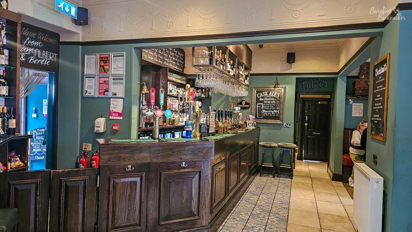 View along the traditional dark wooden bar inside The Albert in Hebden Bridge. Numerous beer hand-pulls and taps line the counter beneath racks of hanging glasses and shelves densely stocked with bottles, while chalkboards advertise cask ales and cocktails. Muted teal walls, dark wood and patterned floor tiles give the room a classic pub appearance, with an open passage continuing beyond the bar towards a black door marked Toilets.
