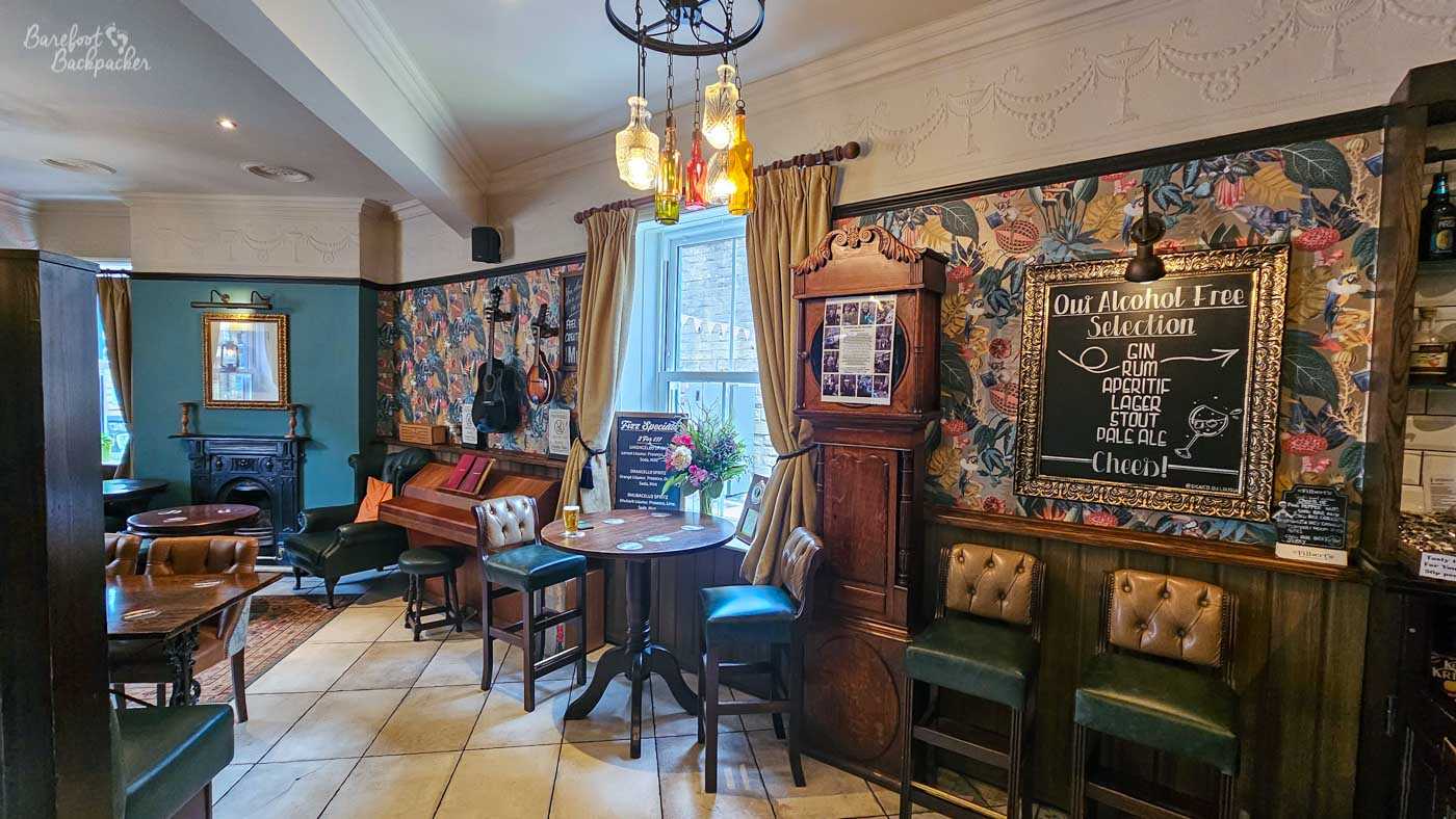 Traditional but colourful interior of The Albert in Hebden Bridge, with dark wood, teal-painted walls and richly patterned botanical wallpaper. Leather and upholstered chairs surround wooden tables, with a small fireplace and an upright piano visible towards the back. A tall wooden grandfather clock stands beside a window, while a large ornate chalkboard advertises the pub’s alcohol-free drinks selection. An unusual chandelier made from illuminated coloured glass bottles hangs overhead.