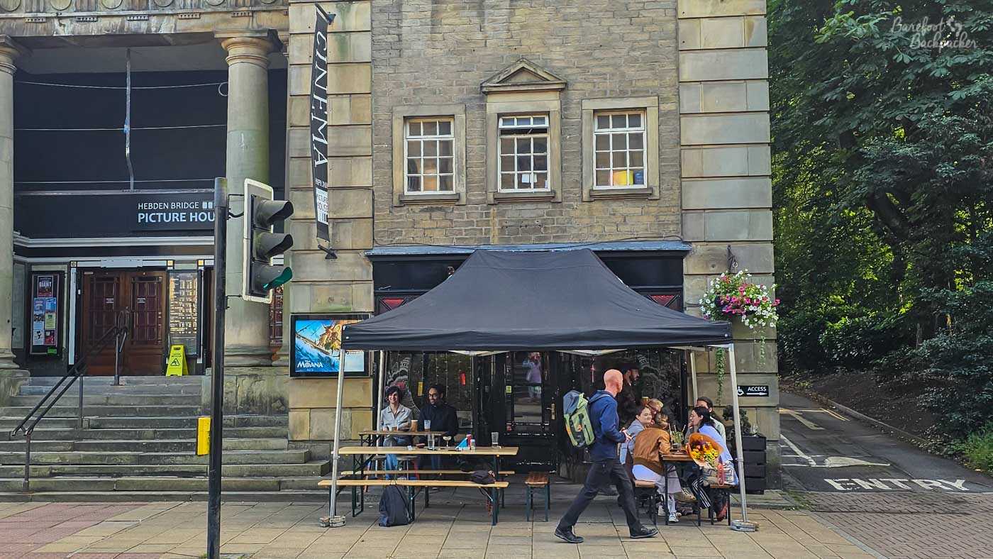 Exterior seating area of Nightjar in central Hebden Bridge, tucked beneath a large black canopy outside a stone building beside Hebden Bridge Picture House. Customers sit around picnic benches and small tables beneath the shelter, while a man with a backpack walks past in the foreground. The bar itself is largely hidden behind the canopy, with no prominent Nightjar sign visible from this angle; trees and a narrow side road border the building on the right.