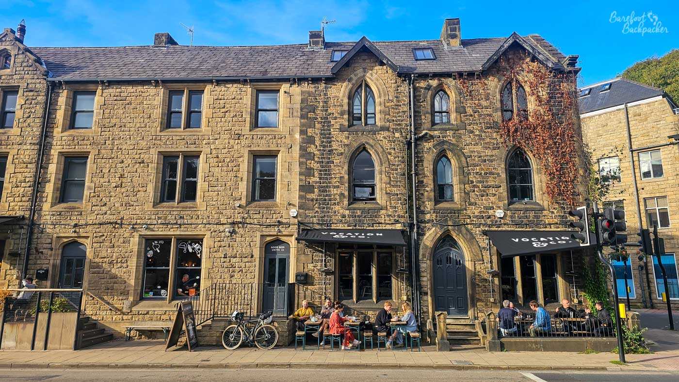 Exterior of Vocation & Co. in Hebden Bridge, occupying part of a handsome multi-storey stone building with striking Gothic-style pointed windows. Two black awnings bearing the Vocation & Co. name mark the entrances, with outdoor tables directly in front where several groups of people are sitting in the sunshine. Bicycles lean against the building nearby, and autumnal climbing foliage covers part of the stonework around the upper windows beneath a vivid blue sky.