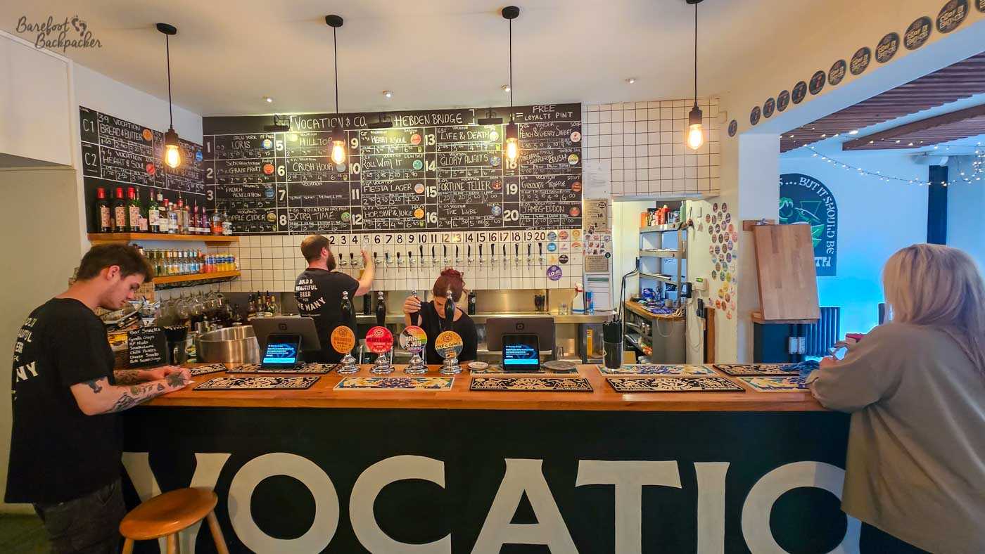 The main bar at Vocation & Co. in Hebden Bridge, with a long wooden counter and staff serving behind it. Twenty numbered beer taps stretch across the white-tiled back wall beneath large handwritten blackboards listing an extensive selection of Vocation and guest beers, including their names, strengths and prices. Bottles and glasses occupy shelves to the left, while large white letters spelling part of “VOCATION” dominate the dark front of the bar.