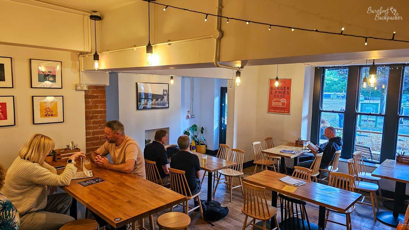 Bright, modern seating area inside Vocation & Co. in Hebden Bridge, with simple wooden tables and spindle-back chairs arranged across a pale-walled room. Several customers sit drinking and talking, while large windows at the far end overlook the street. Exposed brick, framed artwork, hanging bare bulbs and strings of small lights provide decoration, with a poster advertising a burger and fries on one wall. The overall appearance is clean and contemporary compared with a traditional pub interior.
