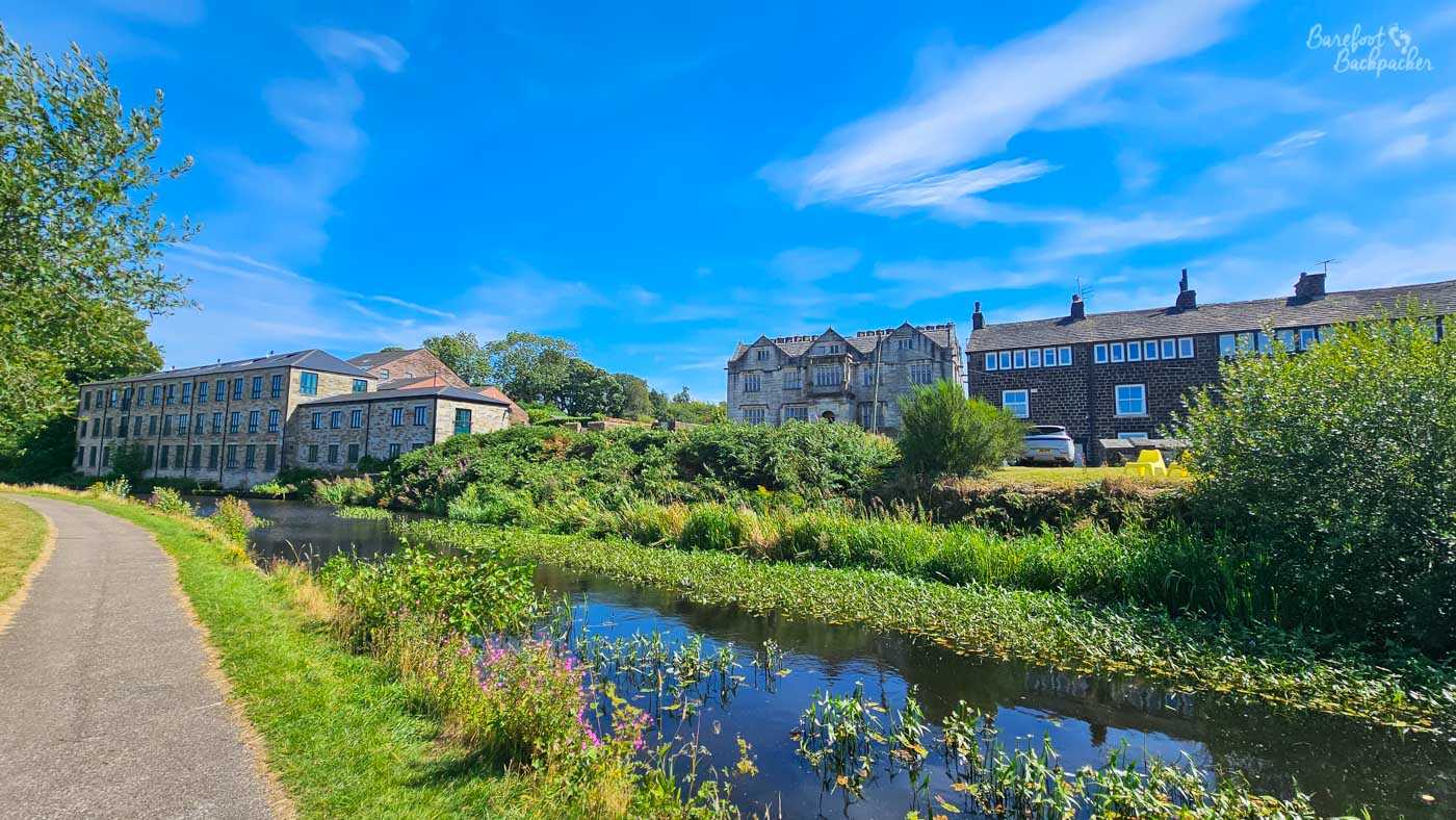 A canal curving past a mixture of substantial old stone buildings and terraced houses beneath a vivid blue sky. Aquatic plants cover parts of the water beside the opposite bank, while a paved towpath curves alongside the canal on the left. Trees, bushes and wildflowers soften the edges between the canal and the buildings.