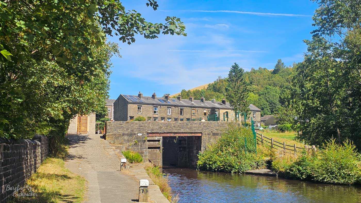 A canal lock surrounded by stone walls and greenery, with the towpath descending towards the lock gates. A row of traditional stone terraced houses stands beyond, backed by trees and the grassy slope of a hill. The scene is partly framed by leafy branches beneath a bright blue summer sky.