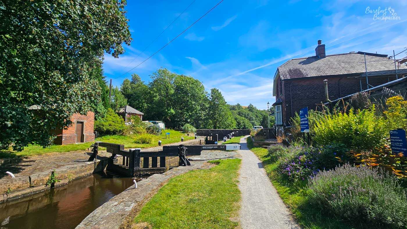 West Summit Lock 37 on the Rochdale Canal, with black-and-white lock gates crossing the water beside the towpath. A stone and brick building stands immediately to the right, surrounded by flowering plants, while trees and smaller buildings fill the background. The lock sits in bright sunshine beneath a deep blue sky.