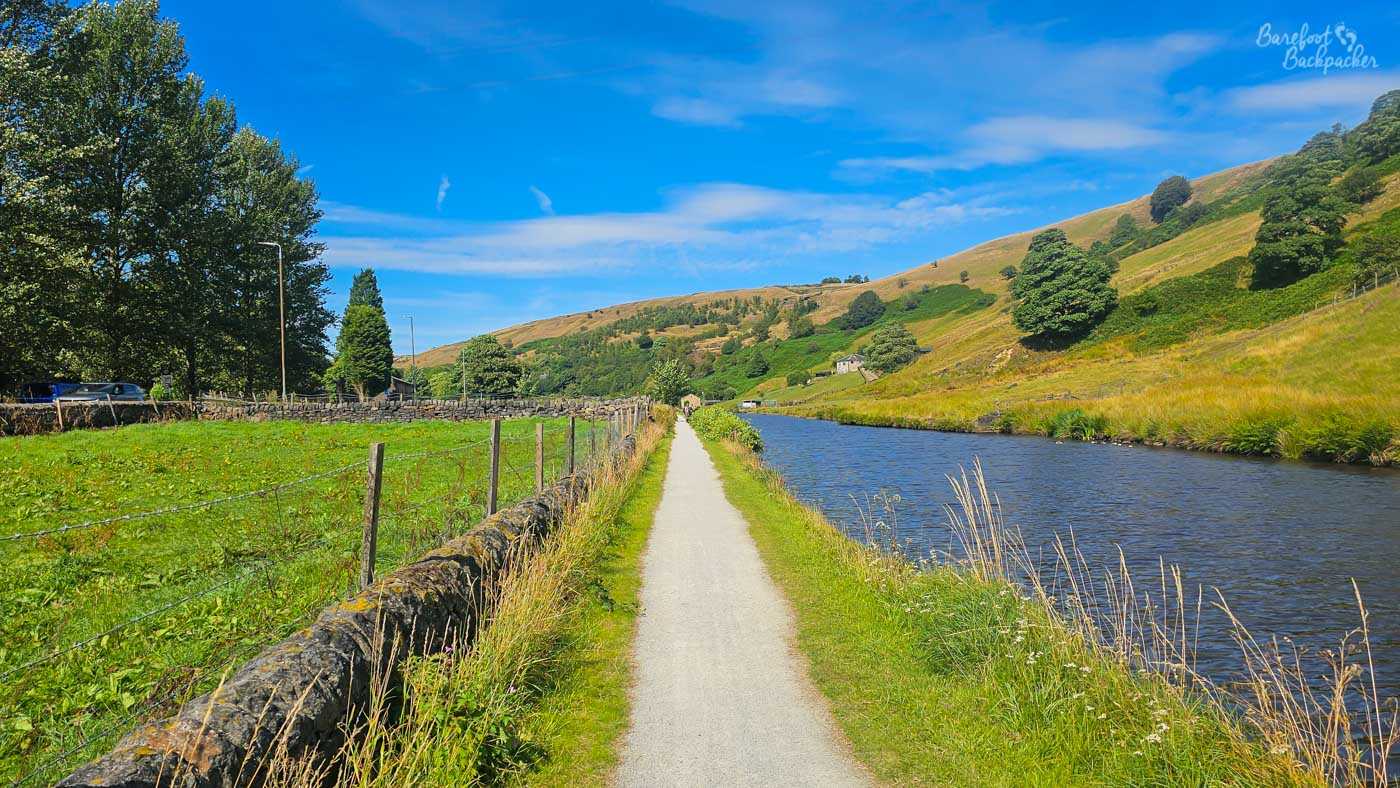 A long straight stretch of canal running through open countryside, with a pale towpath extending into the distance beside the water. A green field and dry-stone wall lie to the left, while steep grassy hills rise from the opposite bank, dotted with trees and a few isolated stone houses. The landscape is brightly lit beneath a clear blue sky streaked with thin white clouds.
