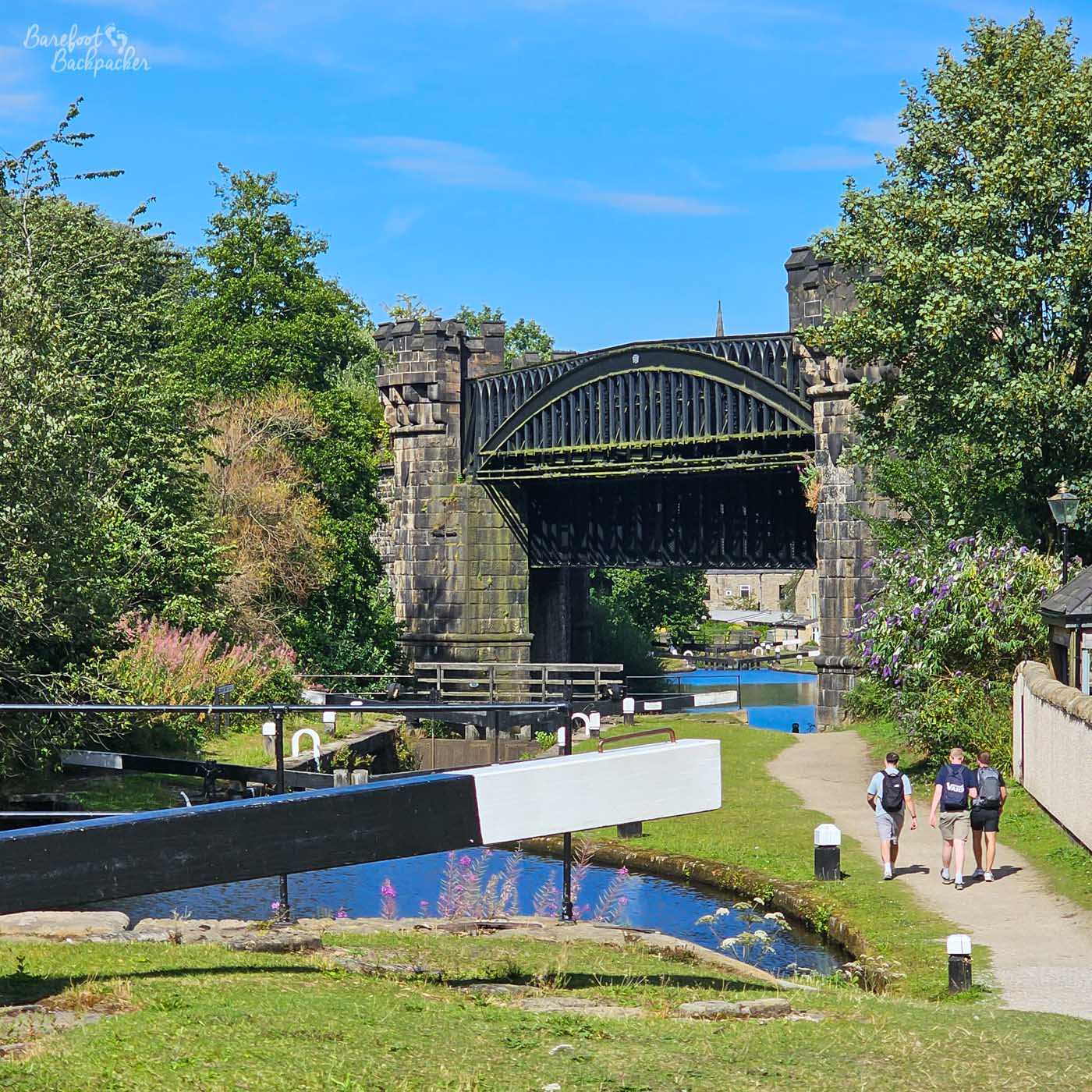 A series of canal locks descending towards a large arched iron bridge supported by substantial stone piers. Black-and-white lock gates cross the foreground, while three people walk along the towpath towards the bridge. Trees and flowering plants surround the canal, with stone buildings visible through the arch beyond.