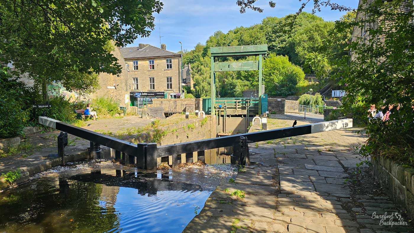 Black-and-white gates at Lock 19 on the Rochdale Canal, viewed from beside the water, with a tall green wooden structure beyond the lock chamber. Stone-paved paths run along both sides, surrounded by trees, bushes and old stone buildings. A few people sit or stand around the lock area in the summer sunshine.