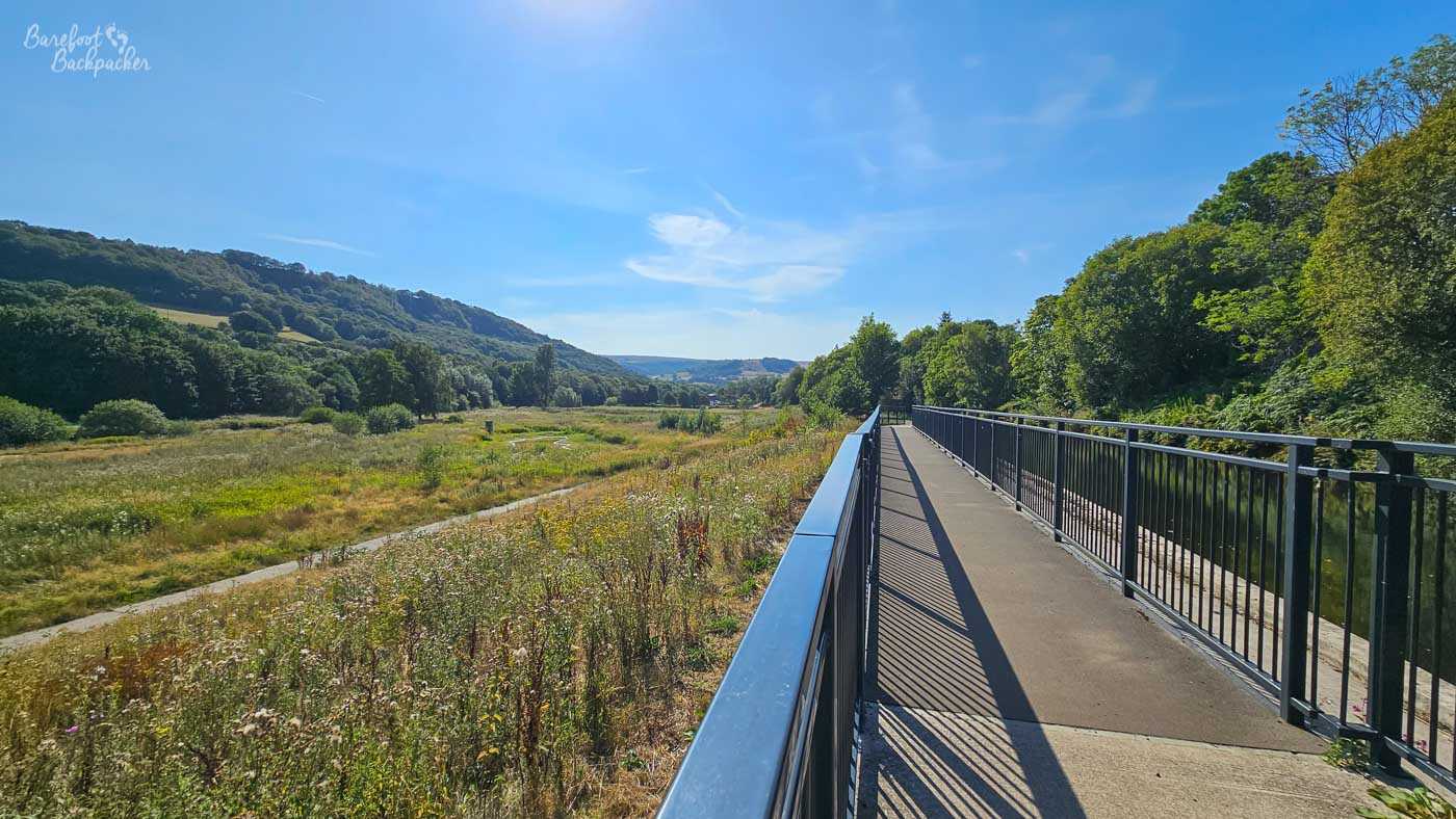 A long, straight pedestrian walkway with black metal railings runs above an open, grassy valley. Wild vegetation and a winding path spread across the broad area to the left, with densely wooded hills rising on either side and more hills visible in the distance. Strong summer sunlight casts long striped shadows from the railings across the walkway.