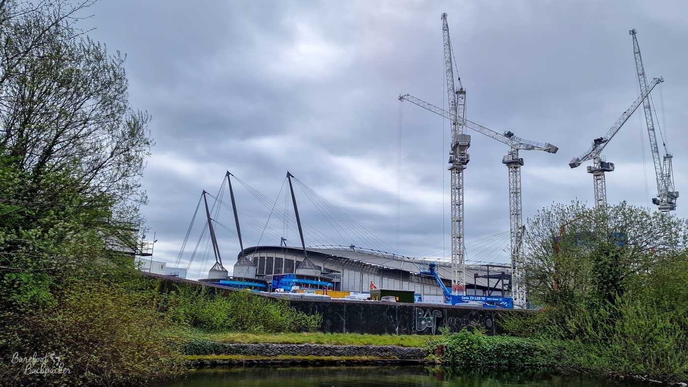 A large modern stadium seen across a canal, its roof supported by tall dark masts and cables. Several enormous construction cranes rise around it against a heavy grey sky, while trees and dense vegetation line the canal bank in the foreground. The dark canal water is just visible along the bottom of the image.