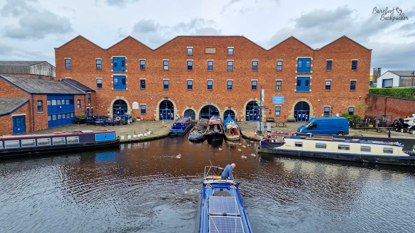 Several narrowboats moored outside a large red-brick former warehouse with distinctive repeated gables, blue-framed windows and arched doorways at water level. More boats are clustered around the basin, while a person stands on the stern of a blue narrowboat in the foreground, surrounded by ripples. The scene has a strongly industrial heritage feel, with the old waterside building dominating the background.
