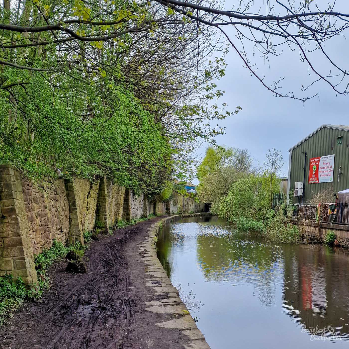 A narrow canal curving gently through a mixture of woodland and industrial buildings beneath an overcast sky. The towpath on the left is heavily churned and muddy, bordered by an old stone retaining wall and overhanging spring foliage. Trees and shrubs crowd the water ahead, while a large grey industrial building stands immediately beside the opposite bank.