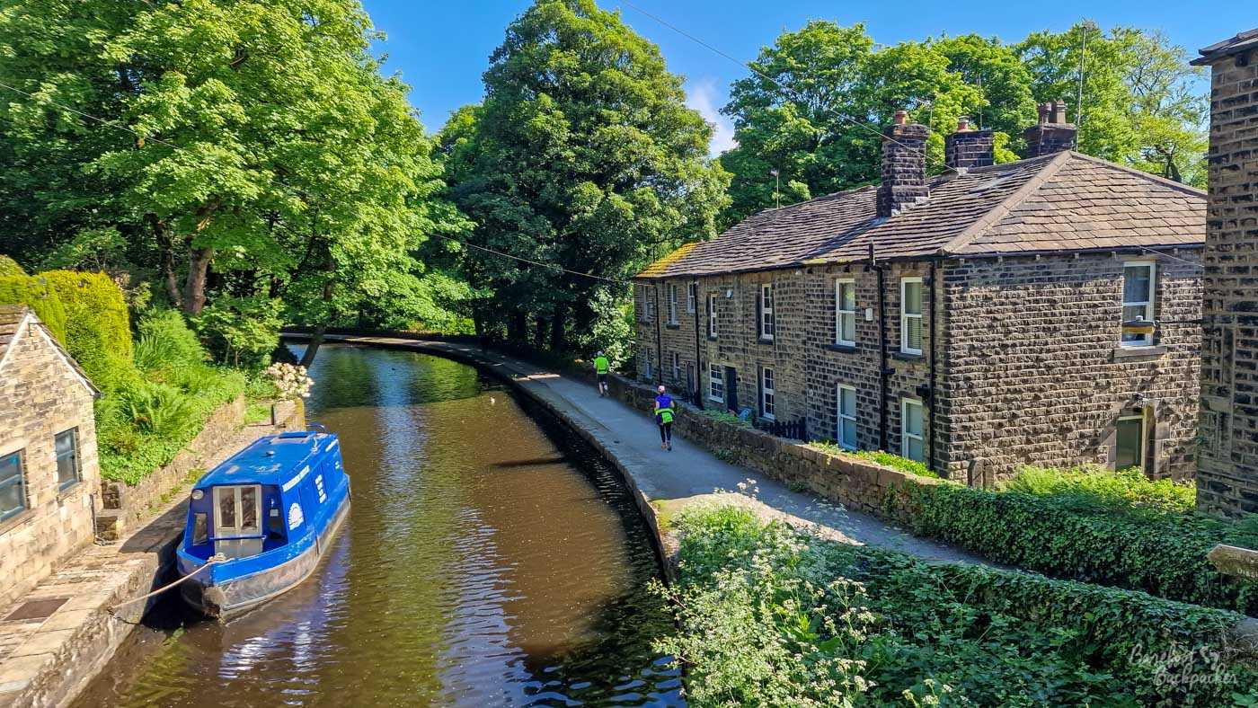 A canal curving between traditional stone cottages and lush green trees on a sunny day. A bright blue narrowboat is moored against the left bank, while two people walk along the towpath beside the houses on the right. The water follows the bend into deep shade beneath the trees.