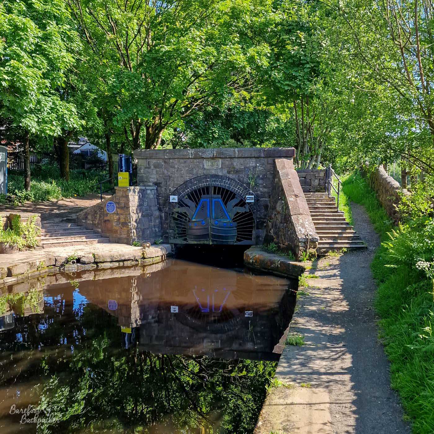 The stone entrance to a canal tunnel surrounded by dense green woodland, with a pair of blue-and-black tunnel gates beneath the arched opening. Stone steps climb on either side of the entrance and a narrow towpath follows the water to the right. The tunnel structure and surrounding trees are reflected in the still, dark water.