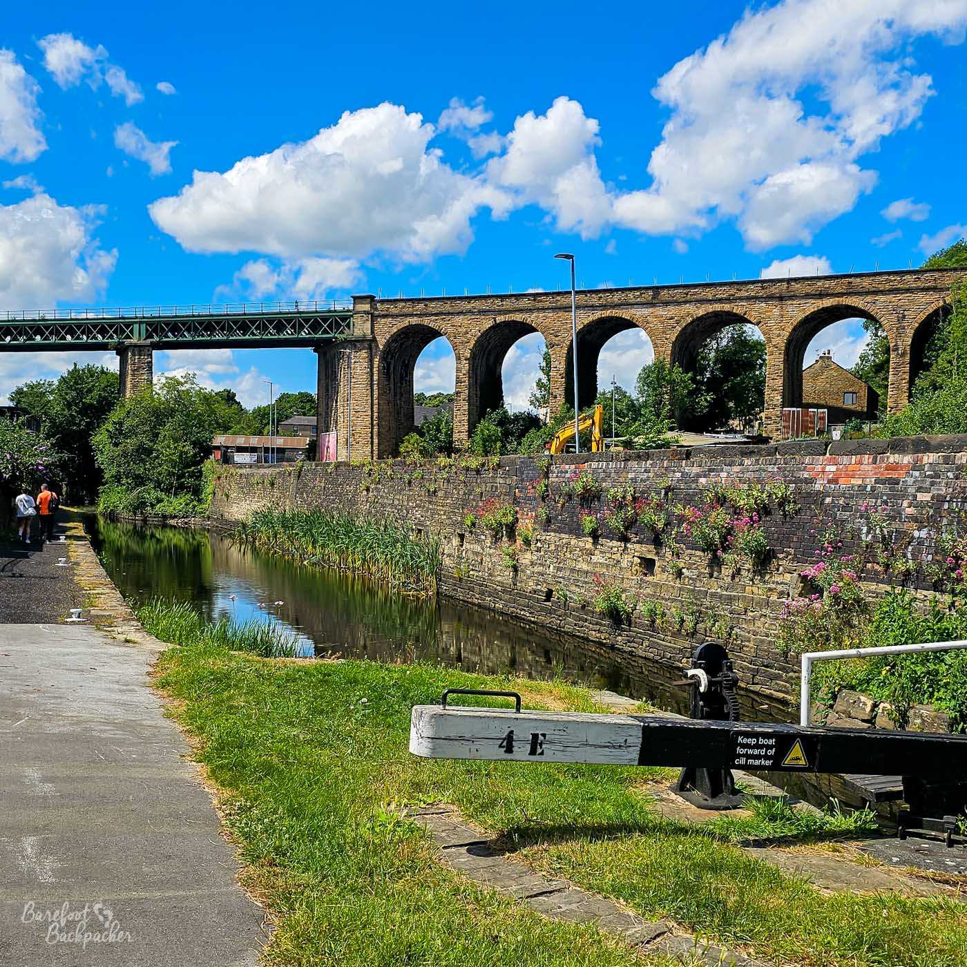 A canal lock beneath a large stone railway viaduct on a bright sunny day. A black-and-white lock beam marked 4E crosses the foreground, while the canal continues alongside a high stone wall dotted with wildflowers. The viaduct's repeated arches stretch across the background, with a separate metal railway bridge joining it on the left.