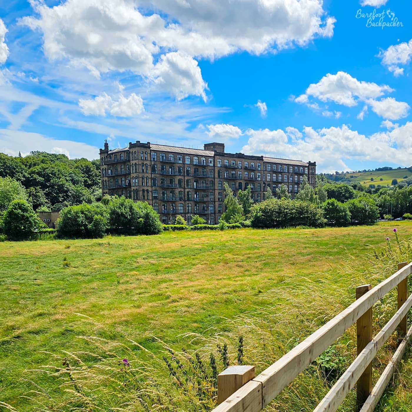 A huge multi-storey stone former mill standing beyond a broad grassy field, surrounded by trees and rolling green hills. Rows of tall windows cover the long industrial building, which dominates the rural landscape. A wooden fence runs through the foreground beneath a vivid blue sky and billowing white clouds.