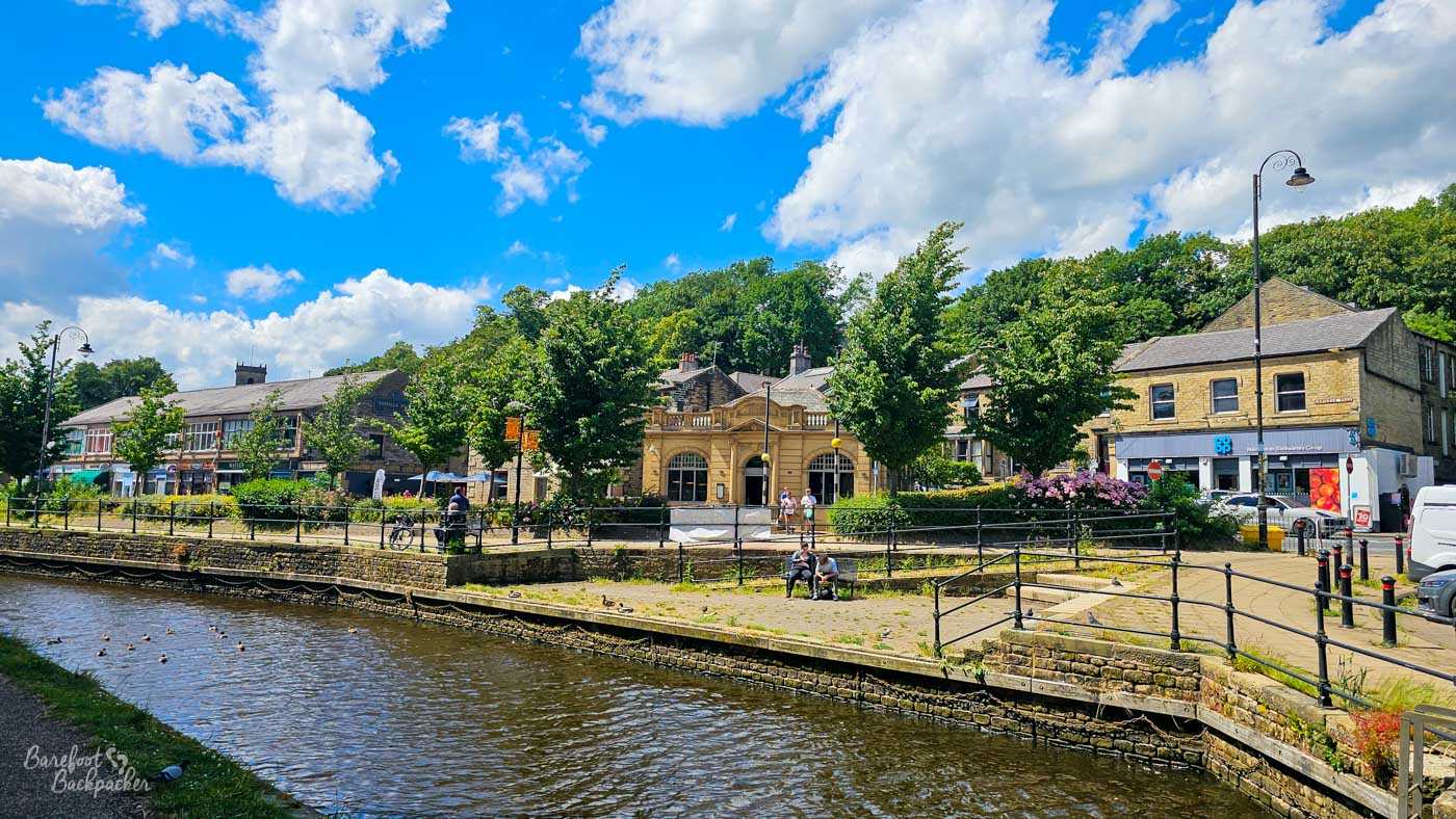 A canal passing through a small town centre, with stone buildings, shops and an ornate historic building lining the opposite bank. People sit and walk beside the water, while ducks are scattered across the canal. Trees, flowers and traditional black railings soften the urban scene beneath a bright blue and cloudy sky.