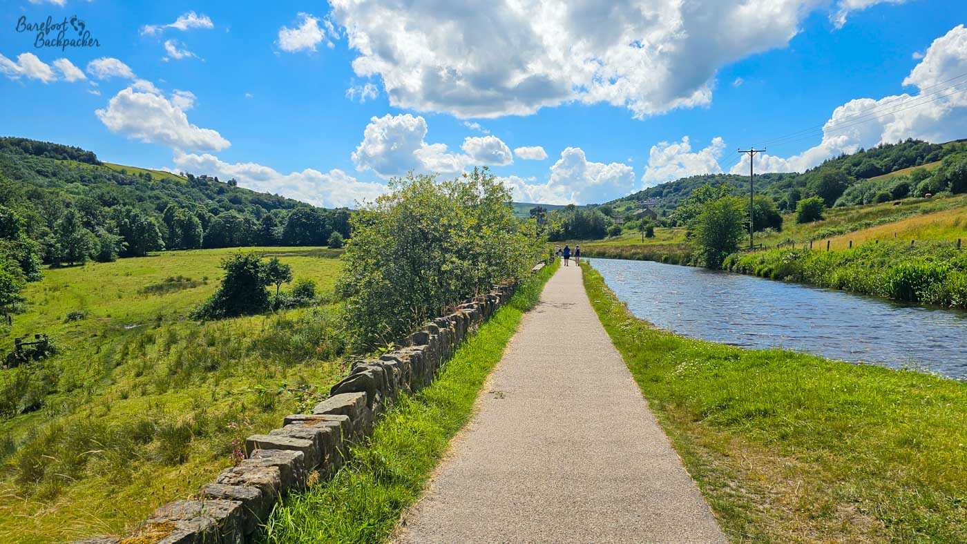 A straight canal and surfaced towpath running through a broad green valley on a bright summer day. A low drystone wall separates the path from fields on the left, while wooded hills rise on either side of the water. Two people walk ahead beneath a vivid blue sky filled with large white clouds.