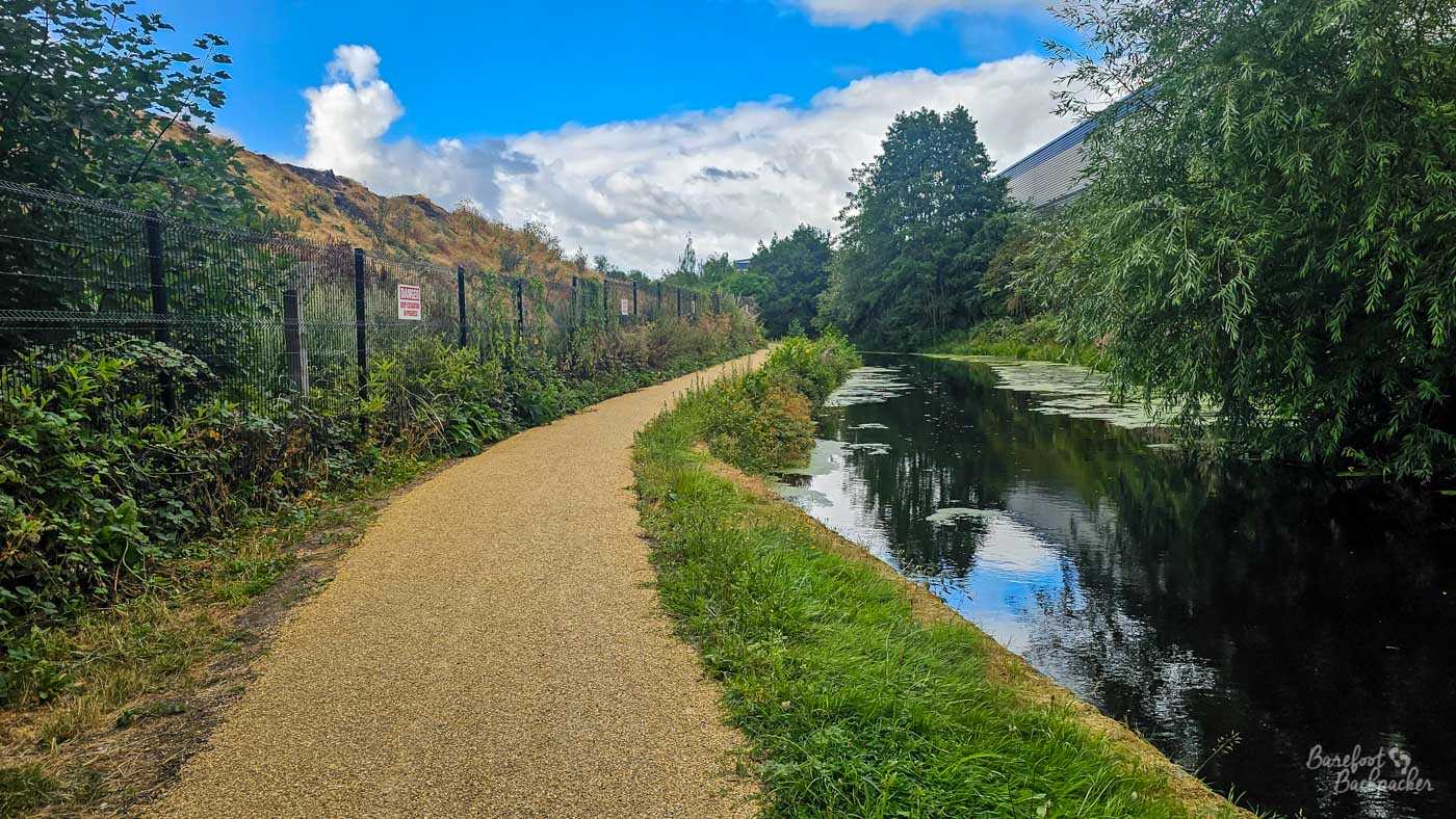 A broad surfaced towpath curving gently alongside a calm canal beneath a bright blue and partly cloudy sky. Trees and shrubs crowd the opposite bank and are reflected in the dark water, with patches of floating vegetation on the surface. A tall security fence follows the left side of the path.