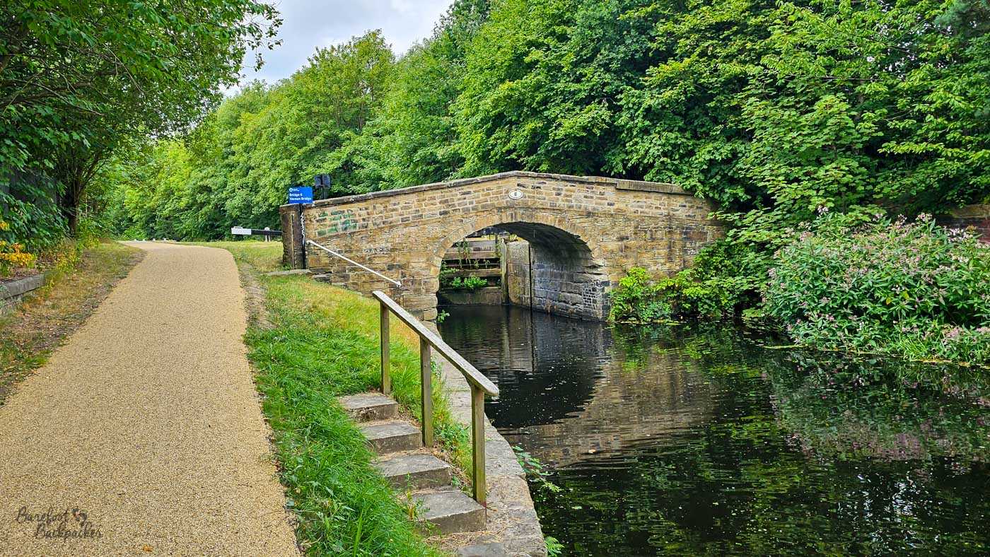A tree-lined canal beside a wide towpath, with a low stone bridge arching over the dark water. Stone steps and a simple wooden handrail descend towards the canal beside the bridge, while lock gates can be seen through the arch. Dense summer greenery surrounds the water and path.