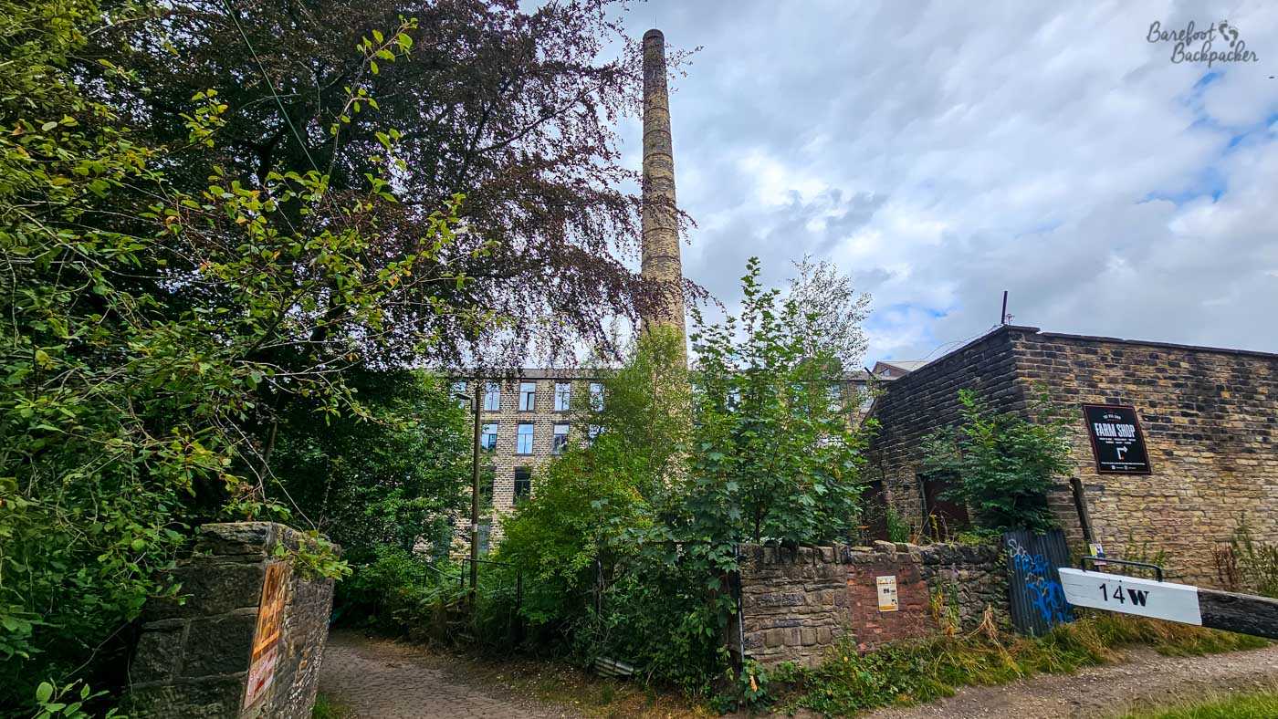 A tall stone industrial chimney rising above trees and old mill buildings beside a canal lock. A black-and-white lock beam marked 14W crosses the lower right of the scene, while dense greenery partly conceals the stone buildings and a path descending between them. The sky is overcast with small patches of blue.