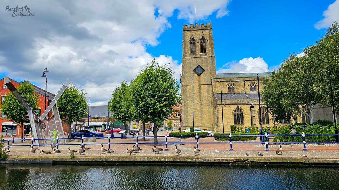 A large sandstone church with a square crenellated tower standing beside a canal in a town centre. Geese and pigeons gather along the paved waterfront behind blue-and-white railings, with shops, roads and a large angular metal sculpture to the left. Dark clouds and brilliant blue sky form a dramatic backdrop above the church.