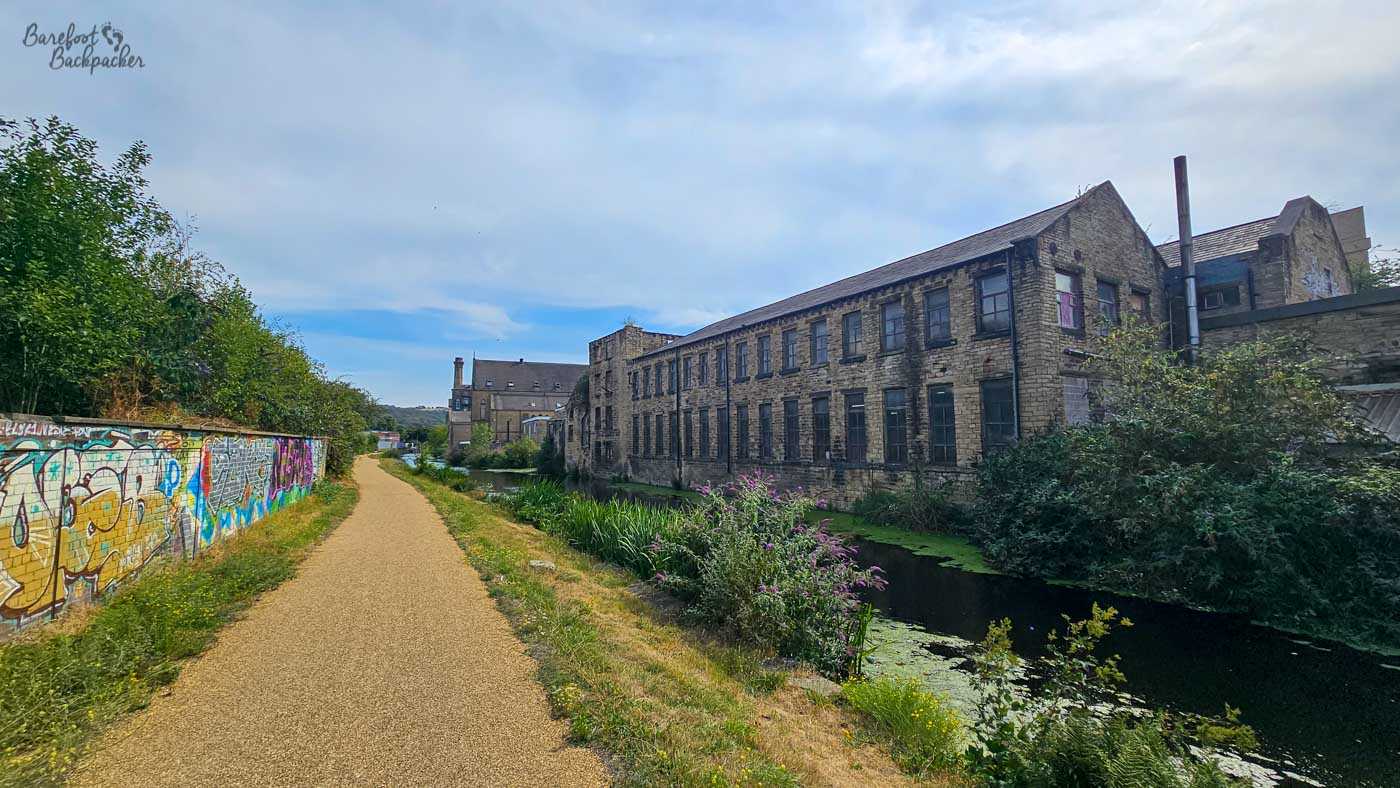 A sunny canal towpath running between a long graffiti-covered wall on the left and an old stone industrial building on the right. The narrow canal is partly hidden by waterside plants and floating greenery, with purple wildflowers growing along its edge. More former industrial buildings stand farther along the route.