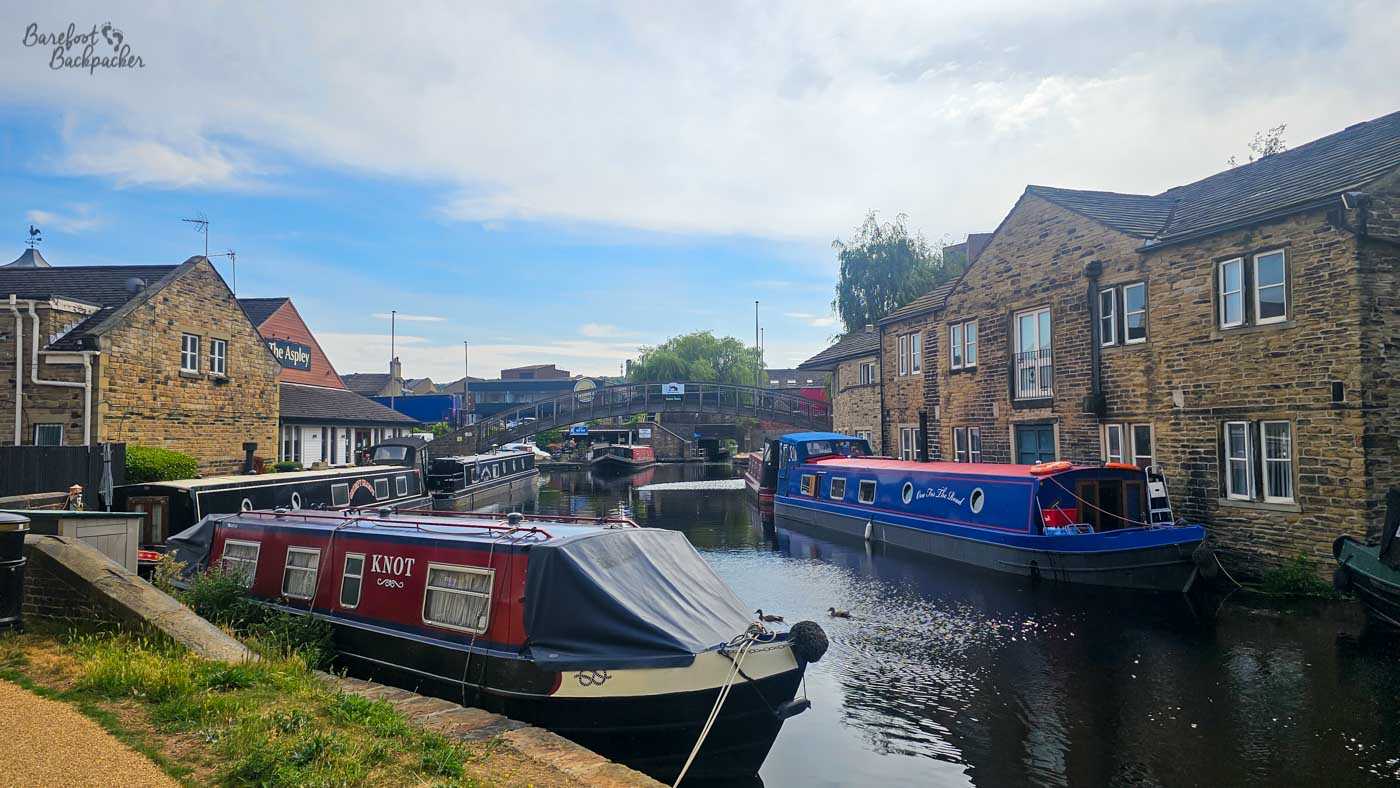 Several colourful narrowboats moored along a canal enclosed by traditional stone buildings. A red-and-black boat named Knot fills the foreground, with blue, black and red boats farther along the water. A pedestrian bridge crosses the canal in the distance beneath a bright blue sky.