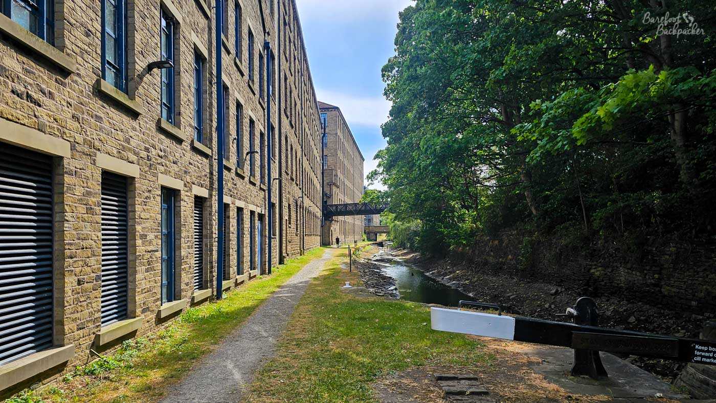A narrow canal channel running between a long stone industrial building on the left and a wooded bank on the right. A rough towpath follows the mill wall towards a small overhead walkway in the distance, while a white-and-black lock beam stretches across the foreground.