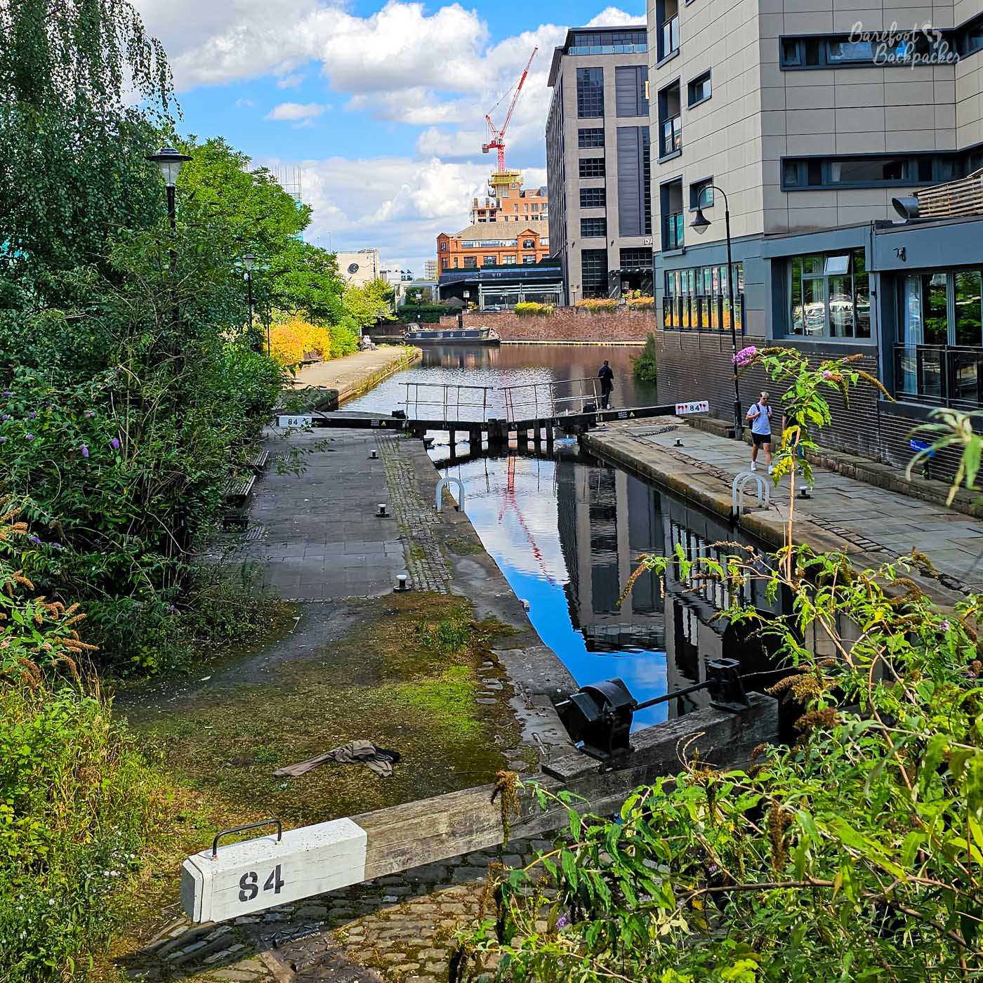 A canal lock running between modern city-centre buildings, viewed from slightly above. Black-and-white lock gates marked 84 cross the water, with paved towpaths on either side and a few people walking nearby. Trees and flowering plants soften the urban surroundings, while a construction crane rises above the buildings in the distance and blue sky is reflected in the still water below.