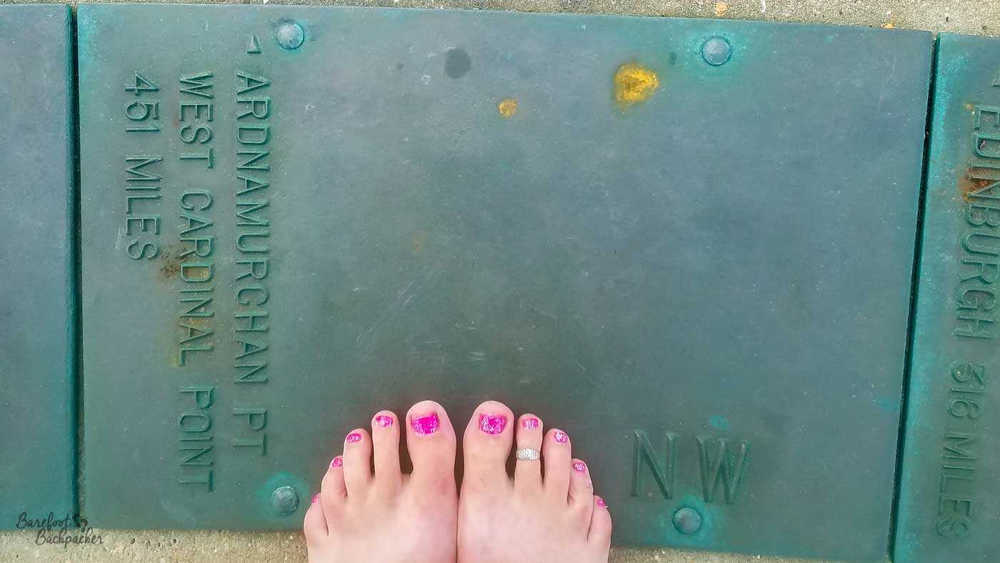 A pair of bare feet with bright pink-painted toenails stand at the bottom of a large green metal direction marker at Ness Point. Embossed lettering points north-west towards Ardnamurchan Point, 451 miles away, with part of another marker for Edinburgh visible at the right edge.