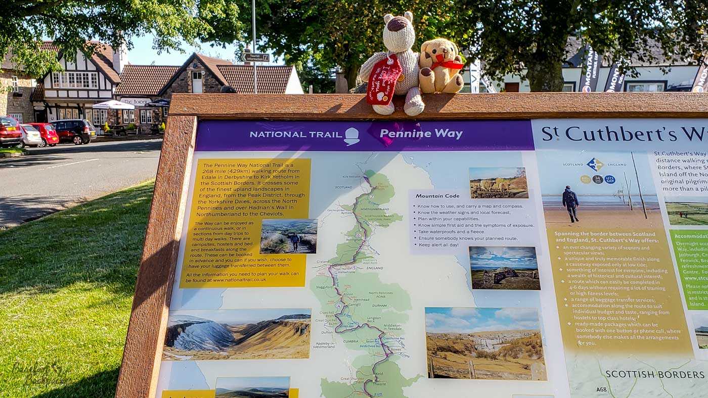 This image shows a signpost detailing the Pennine Way National Trail and St. Cuthbert's Way. The sign includes a map of the route, showing its path and provides practical information for walkers, such as the importance of knowing how to use a map and compass, checking weather forecasts, and carrying essential safety items. Nearby, St. Cuthbert's Way is described as a 100-mile walking route that spans the border between Scotland and England, offering diverse scenery and historical points of interest. The sign is situated outdoors, with trees and buildings visible in the background, and two teddy bears are placed on top of the sign as a whimsical touch.