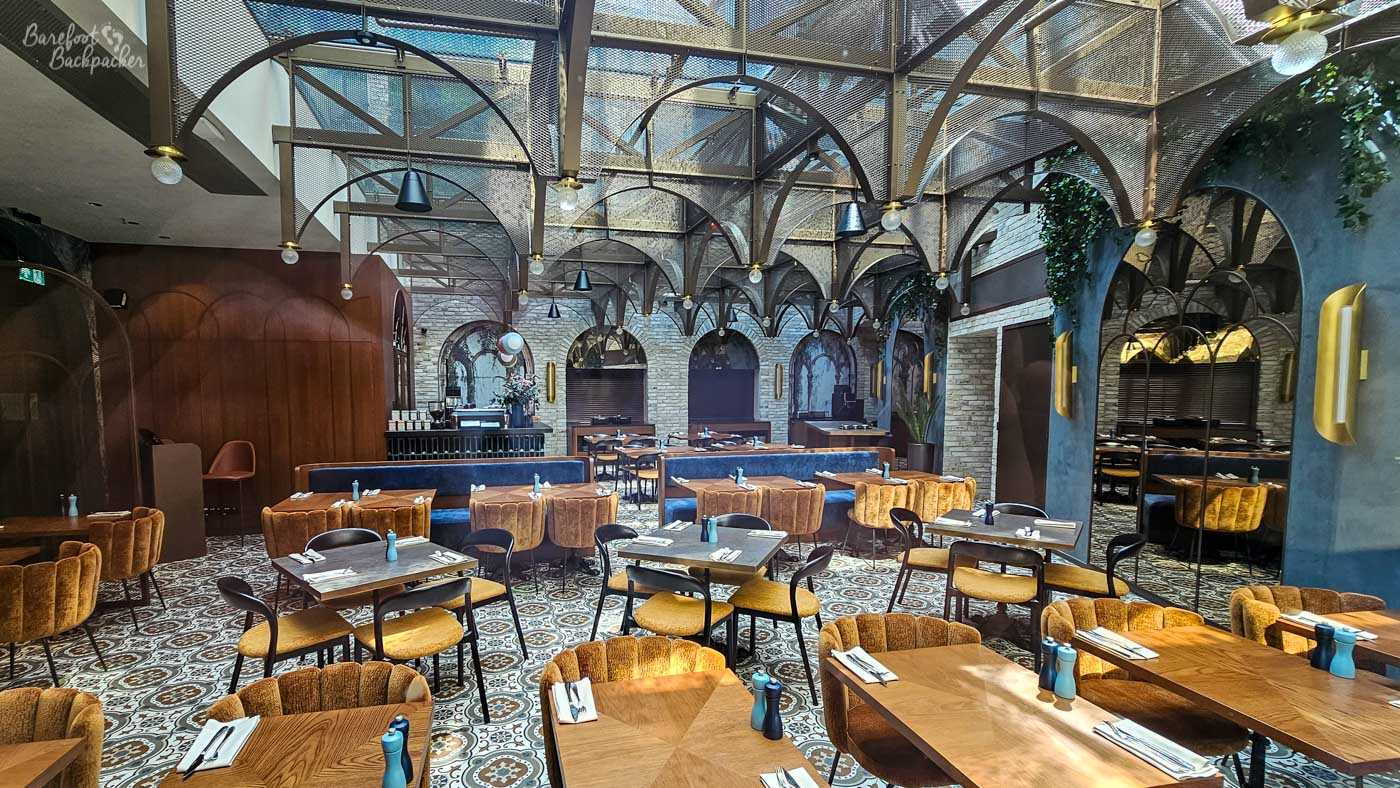 The stylish interior of a modern restaurant in Kraków, with wooden tables surrounded by mustard-coloured upholstered chairs and curved blue banquette seating. Decorative patterned floor tiles run throughout the room, while dark metal arches and mesh panels form an elaborate vaulted framework overhead, echoed by large arched mirrors along the walls. Warm sunlight falls across several of the tables, contrasting with the darker wood, exposed pale brickwork and industrial-style detailing elsewhere in the dining room..