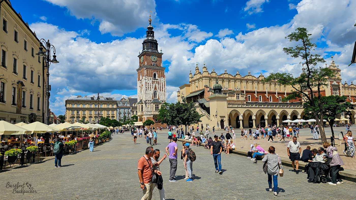 A busy view across Kraków's Main Market Square, with the tall brick Town Hall Tower rising prominently on the left and the long arcaded Cloth Hall stretching across the right. Outdoor cafés occupy the left side of the square while tourists walk, gather and sit around the broad pedestrian space beneath a vivid blue sky filled with dramatic white clouds. The combination of Gothic tower, Renaissance architecture and bustling public space gives a wide overview of the historic centre.