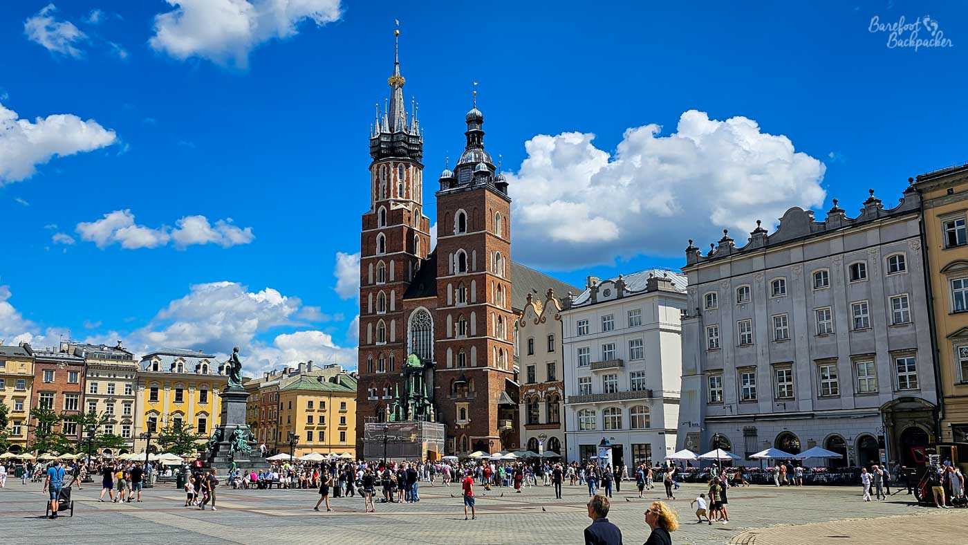 St Mary's Basilica dominating Kraków's busy Main Market Square, its two unequal red-brick towers rising dramatically against a deep blue sky and huge white summer clouds. Colourful historic townhouses line the square on either side, while the Adam Mickiewicz Monument stands to the left of the church. Crowds of visitors fill the large paved square below, giving the scene a lively summer atmosphere.