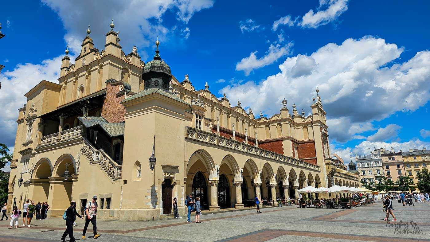 The historic Cloth Hall, or Sukiennice, on Kraków's Main Market Square beneath a spectacular blue sky filled with large white clouds. The long cream-coloured Renaissance building has an ornate roofline, a row of pointed arches along its ground floor and an external staircase rising to an upper gallery at the near end. Visitors cross the broad square outside, with café tables and umbrellas lining part of the arcade.