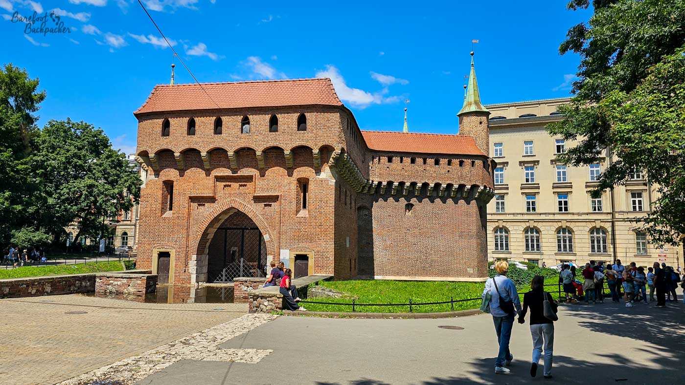 The Barbican of Kraków, a substantial medieval red-brick fortification standing in bright summer sunshine beneath a vivid blue sky. The defensive structure has a deep pointed entrance arch, rows of narrow openings and projecting brickwork beneath its upper walls, with red tiled roofs and small green-topped turrets. People walk and sit around the entrance, while leafy trees frame the historic building on either side.