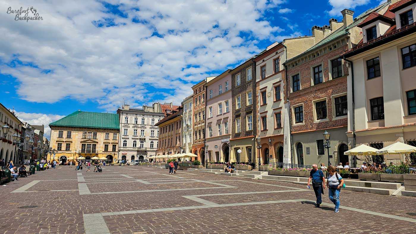 A broad, sunny cobbled square in Kraków's historic centre, lined on the right by a varied row of colourful old townhouses in brick, cream, pink and other muted shades. Café terraces with pale umbrellas run beneath the buildings, while scattered pedestrians cross the large open space. Bright blue sky and dramatic broken white clouds make the square feel particularly open and summery.