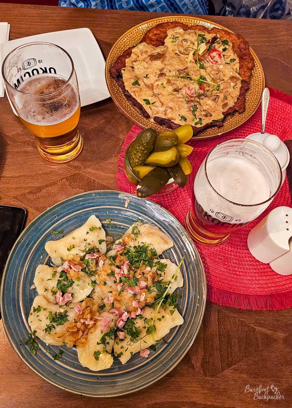 A table laden with Polish food and beer, including a large blue plate of pierogi topped with fried onions, small pieces of bacon and chopped fresh herbs. Behind them is a large potato pancake covered in a creamy mushroom sauce and garnished with herbs and halved cherry tomatoes, with a small dish piled with pickled cucumbers between the two meals. Two partly drunk glasses of pale beer complete the extremely substantial spread.