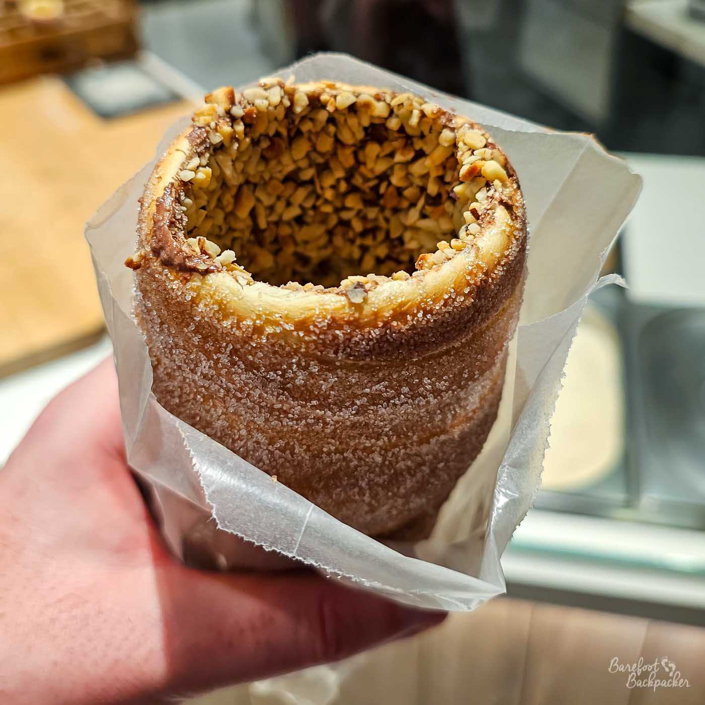 A freshly baked cylindrical chimney cake held in a paper wrapper, its crisp golden-brown exterior coated with sugar and its hollow interior thickly covered in chopped nuts. The close-up view looks directly into the open top of the pastry, showing its spiral layers and generous nut coating. A hand supports it from below while the bakery counter behind is softly out of focus.