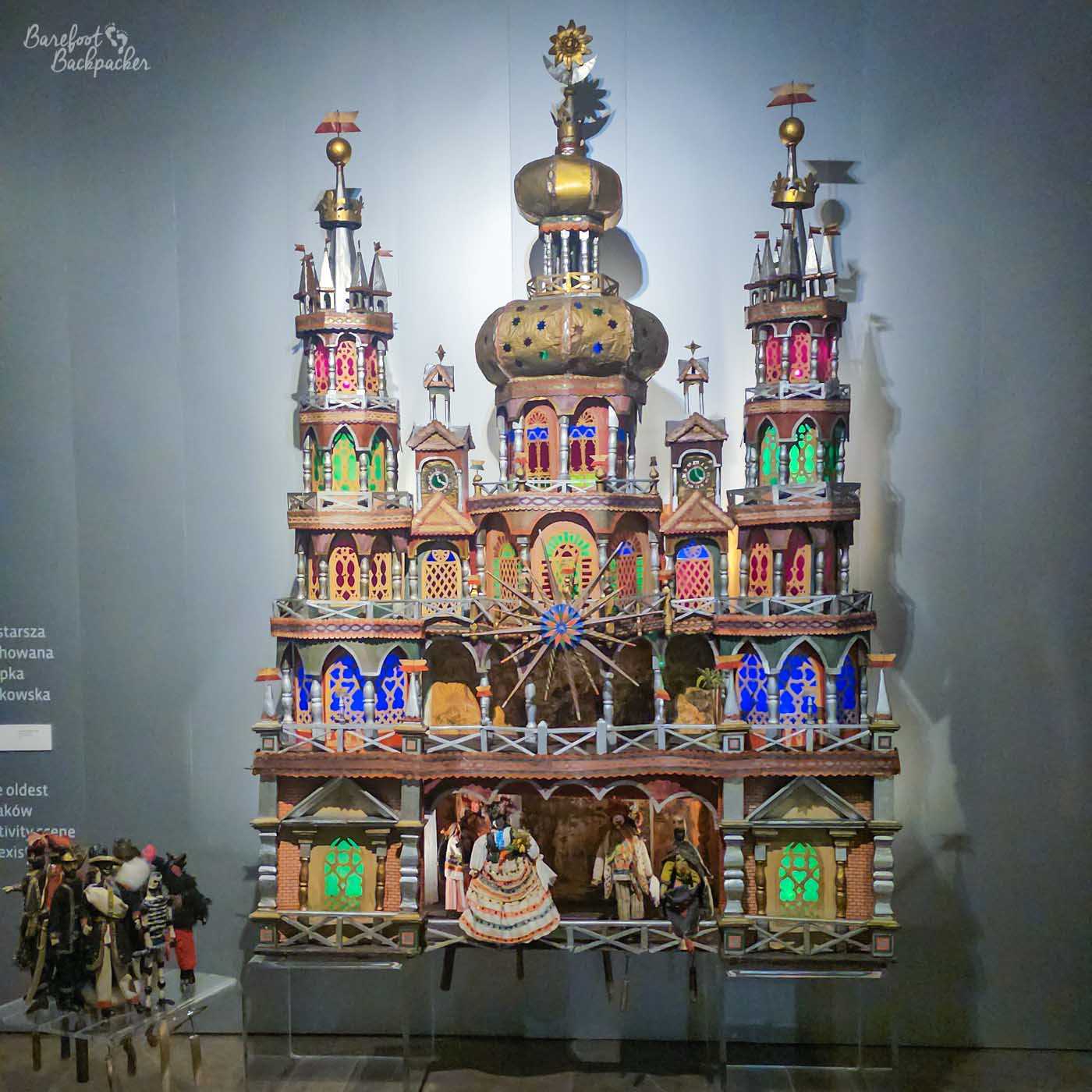 An elaborate traditional Kraków nativity scene, or szopka krakowska, displayed against a plain grey wall. The tall, fantastical structure resembles a brightly coloured miniature palace or cathedral, with multiple towers, spires, domes and tiny arched windows picked out in vivid reds, greens, blues and gold. Small figures in traditional dress appear on the lower level, while a large decorative starburst occupies the centre of the intricate, highly symmetrical model.