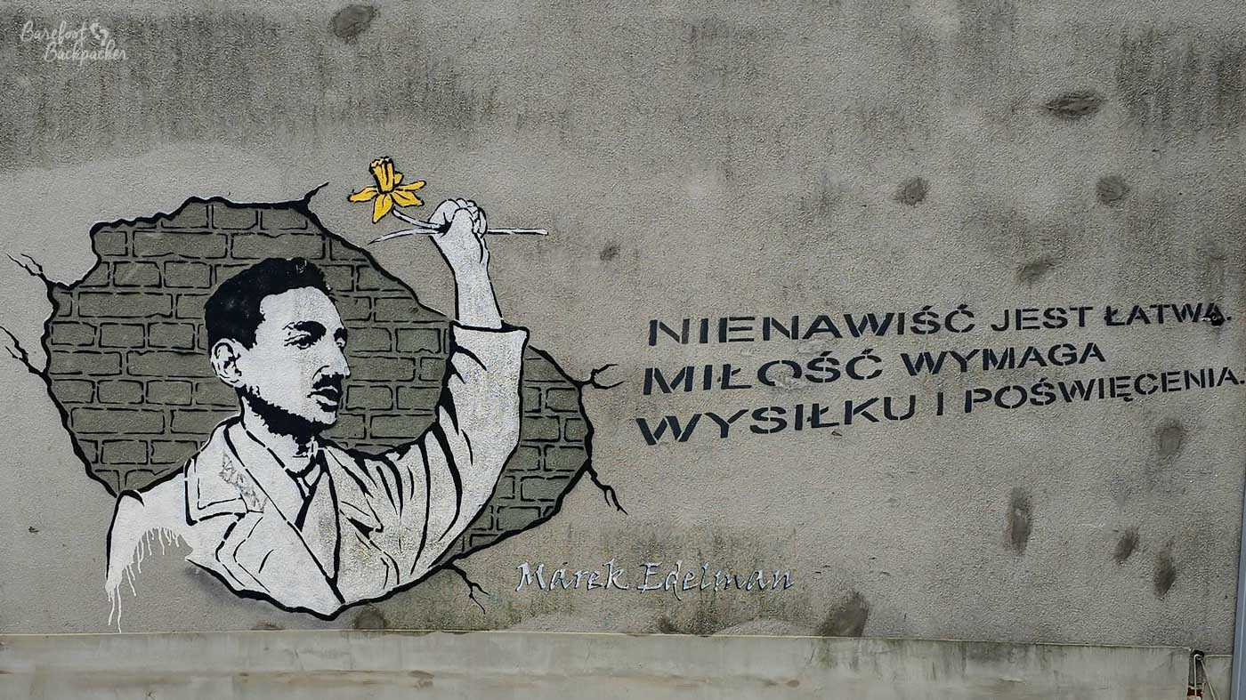 A black, white and yellow mural of Marek Edelman painted onto a weathered grey wall in Kraków. Edelman is depicted raising one arm and holding a small yellow flower, apparently emerging through a broken section of painted brickwork. Alongside him is the Polish quotation: Nienawiść jest łatwa. Miłość wymaga wysiłku i poświęcenia, meaning approximately: Hatred is easy. Love requires effort and sacrifice.