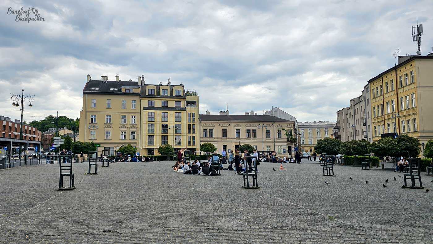 A broad cobbled square in Kraków's Kazimierz district, surrounded by pale yellow and cream buildings beneath a heavy grey sky. Groups of people sit informally on the paving around the centre of the square, while others gather near the buildings and pigeons wander across the open foreground. Sparse young trees in protective frames dot the otherwise largely empty expanse of cobbles. The square is also filled with large, human-sized, chair frames.