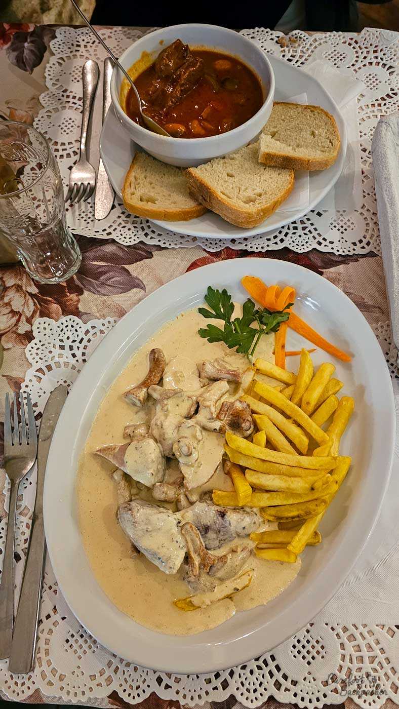 A large plate of meat and mushrooms in a rich creamy sauce, served with a generous pile of chips and a small garnish of parsley and sliced carrot. Behind it is a bowl of deep reddish-brown stew or soup, accompanied by three thick slices of bread. The closely framed overhead photograph captures the hearty scale of the meal, served on white plates over decorative lace-patterned table coverings.