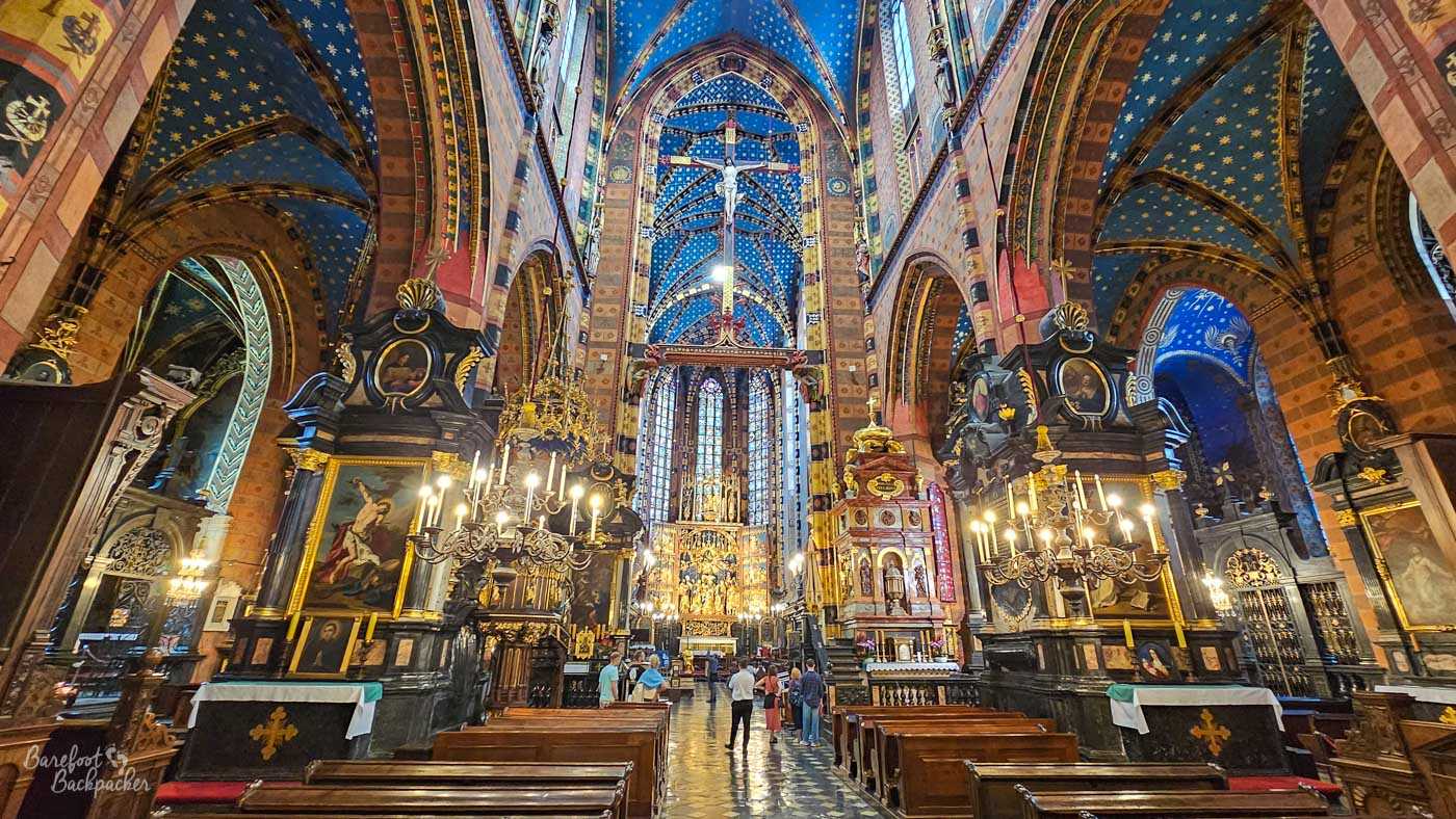 The richly decorated interior of St Mary's Basilica in Kraków, looking along the nave towards the altar. Tall red and patterned columns rise into deep blue vaulted ceilings scattered with golden stars, while ornate dark side altars, paintings, gilded decoration and large chandeliers fill almost every part of the interior. A monumental crucifix hangs high above the central aisle, with visitors standing beneath it and exploring the church.
